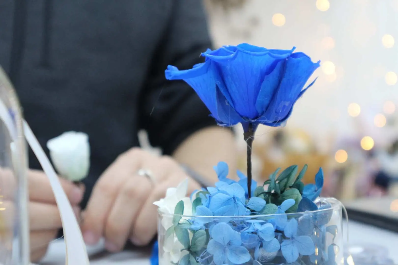 Close-up of Myosotis.flower Kwun Tong preserved flower DIY workshop, showing a participant intently arranging a blue preserved rose and small white flowers into a glass container.