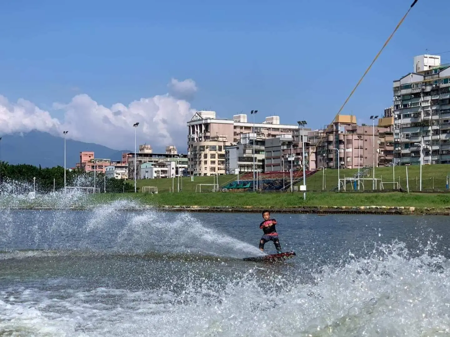 A wakeboarder rapidly glides across Taipei's Breeze Canal, creating an impressive spray, highlighting the speed and excitement of this extreme water sport with urban buildings in the background.