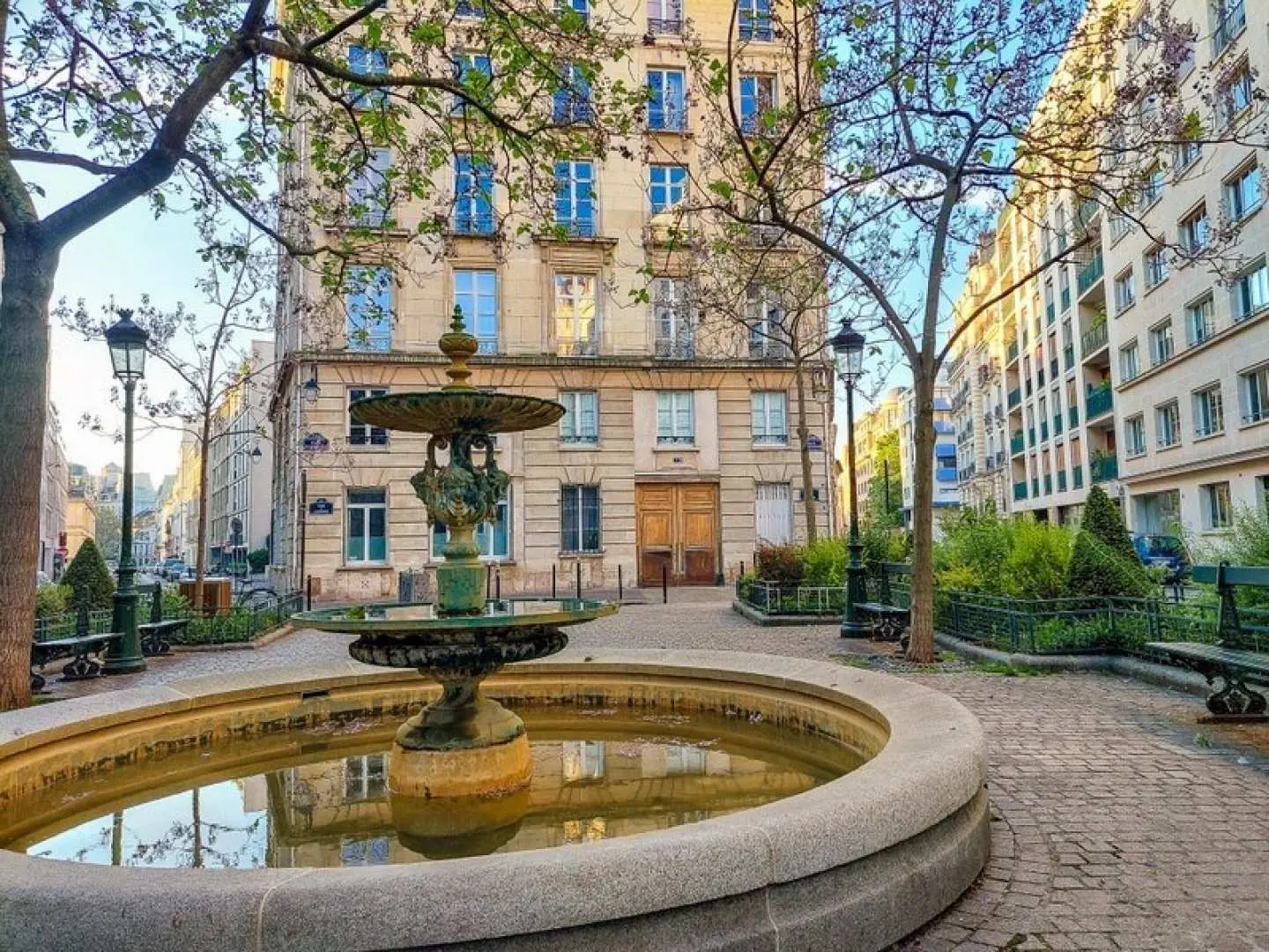 A charming public square in central Paris featuring a beautiful fountain surrounded by classic buildings and trees, an iconic filming location near Emily's apartment on the 'Emily in Paris' walking tour.