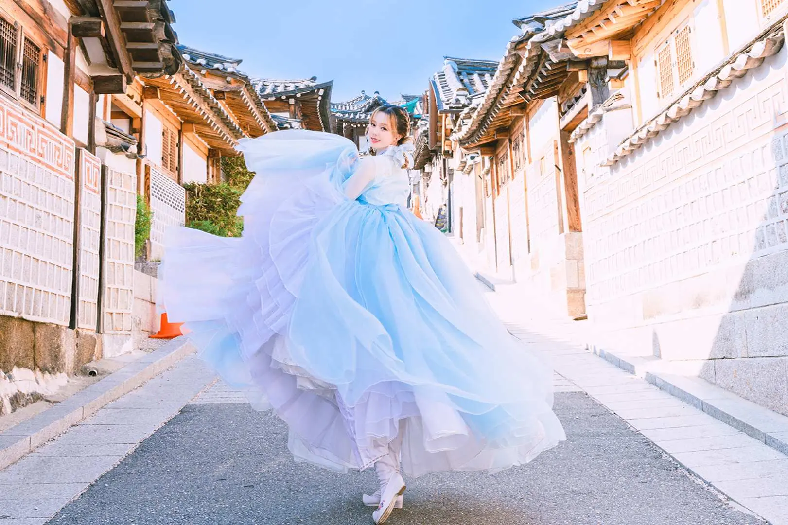A woman joyfully twirling in an elegant light blue and purple gradient Hanbok on a picturesque traditional street in Seoul, capturing dynamic moments of the outdoor photoshoot.