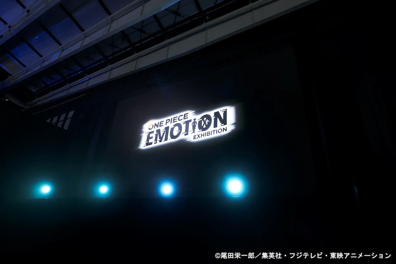 โลโก้ 'ONE PIECE EMOTION EXHIBITION' ที่เปล่งแสงในห้องมืด พร้อมไฟสปอร์ตไลต์ สร้างบรรยากาศทางเข้าที่น่าตื่นตาตื่นใจและดื่มด่ำในงานโอซาก้า