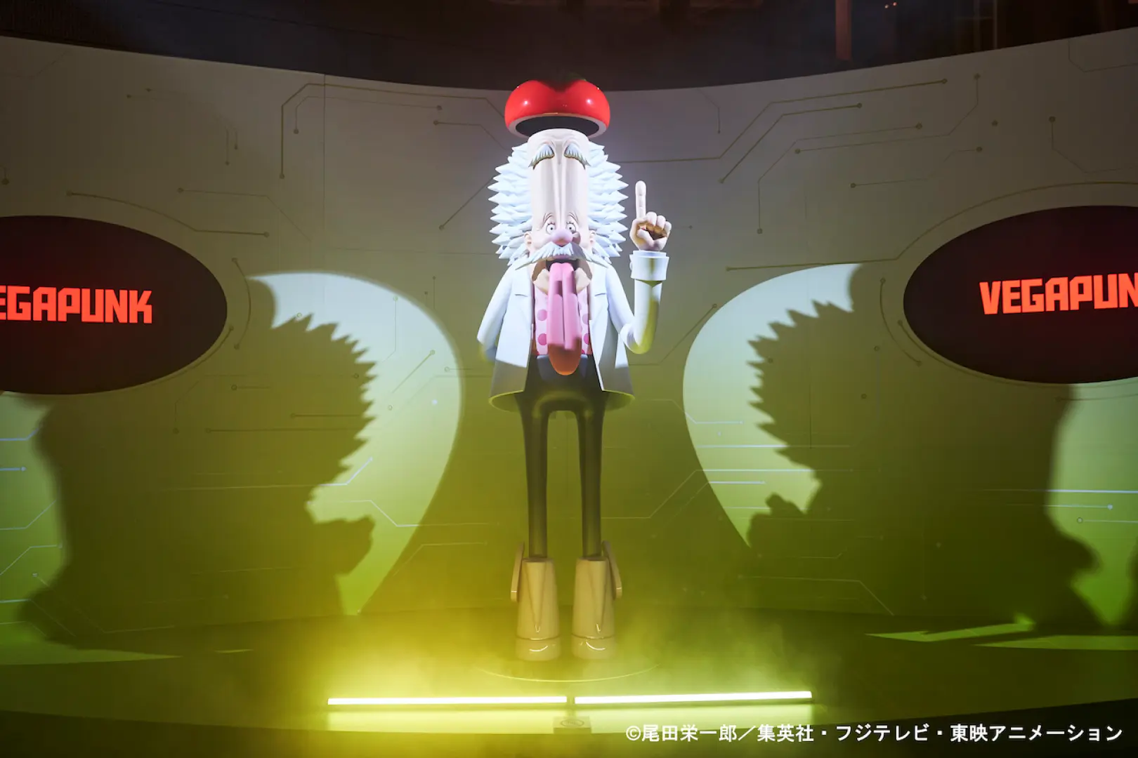 รูปปั้นขนาดเท่าตัวจริงของดอกเตอร์เวก้าพังค์จากภาคเอ็กเฮดของ 'ONE PIECE' โดดเด่นด้วยสมองและเสื้อกาวน์ที่เป็นเอกลักษณ์ สว่างไสวด้วยแสงสีเขียวที่นิทรรศการโอซาก้า