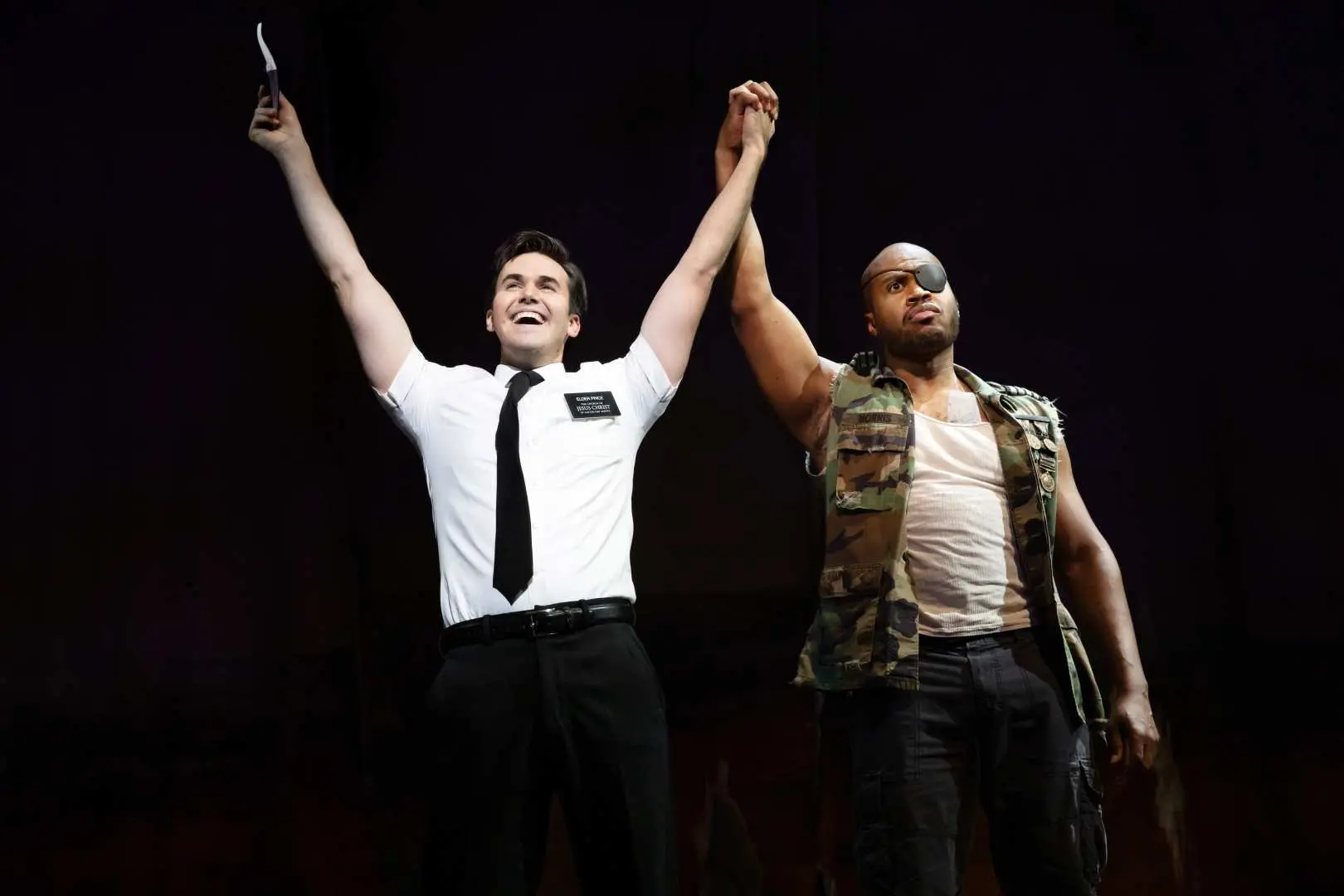 Curtain call photo from 'The Book of Mormon' Broadway musical, with two main actors raising their hands; one missionary holds a knife, conveying the show's humor and dramatic tension.