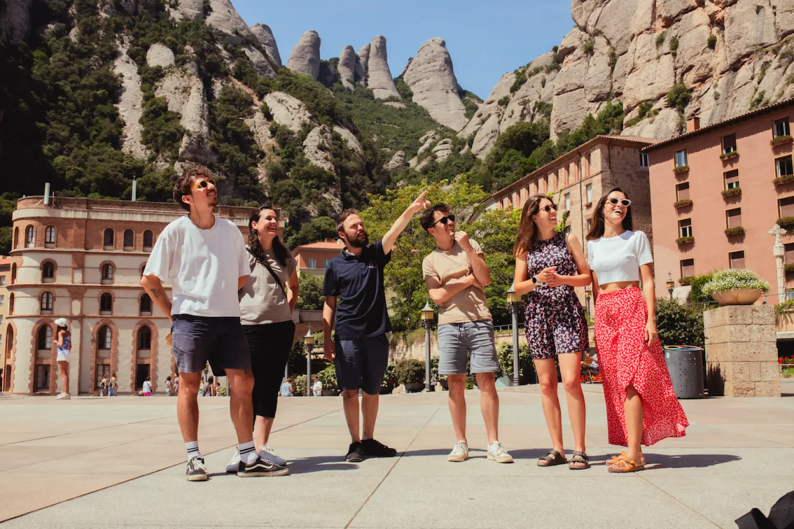 A guide pointing out the unique jagged Montserrat mountain peaks to tourists, with the magnificent monastery buildings in the background, during a half-day tour.