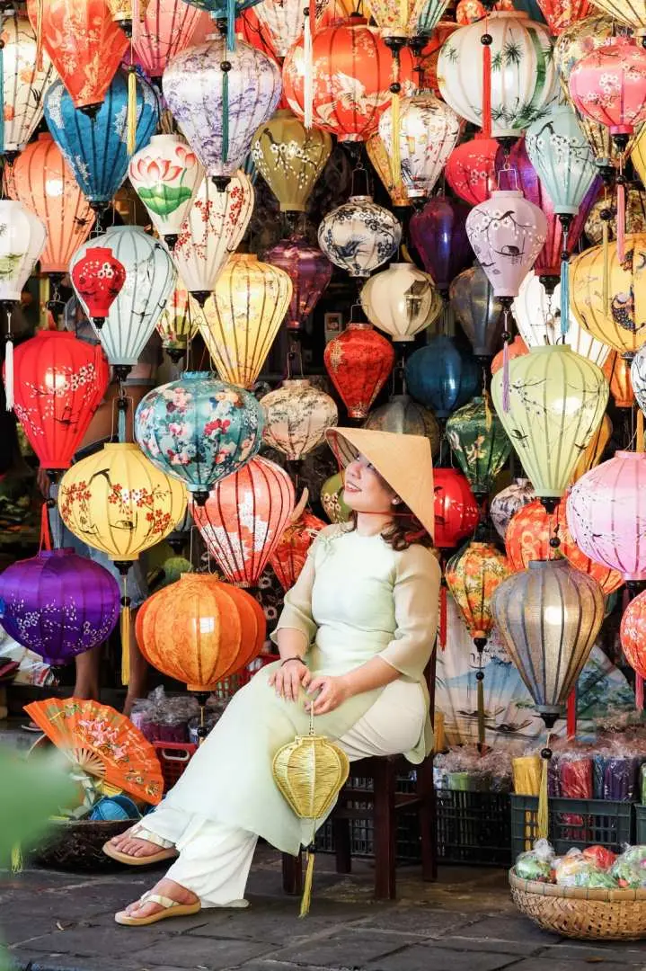 A woman in traditional Vietnamese attire elegantly sitting amidst a vibrant display of colorful silk lanterns in an artistic lantern shop within Hoi An Ancient Town.