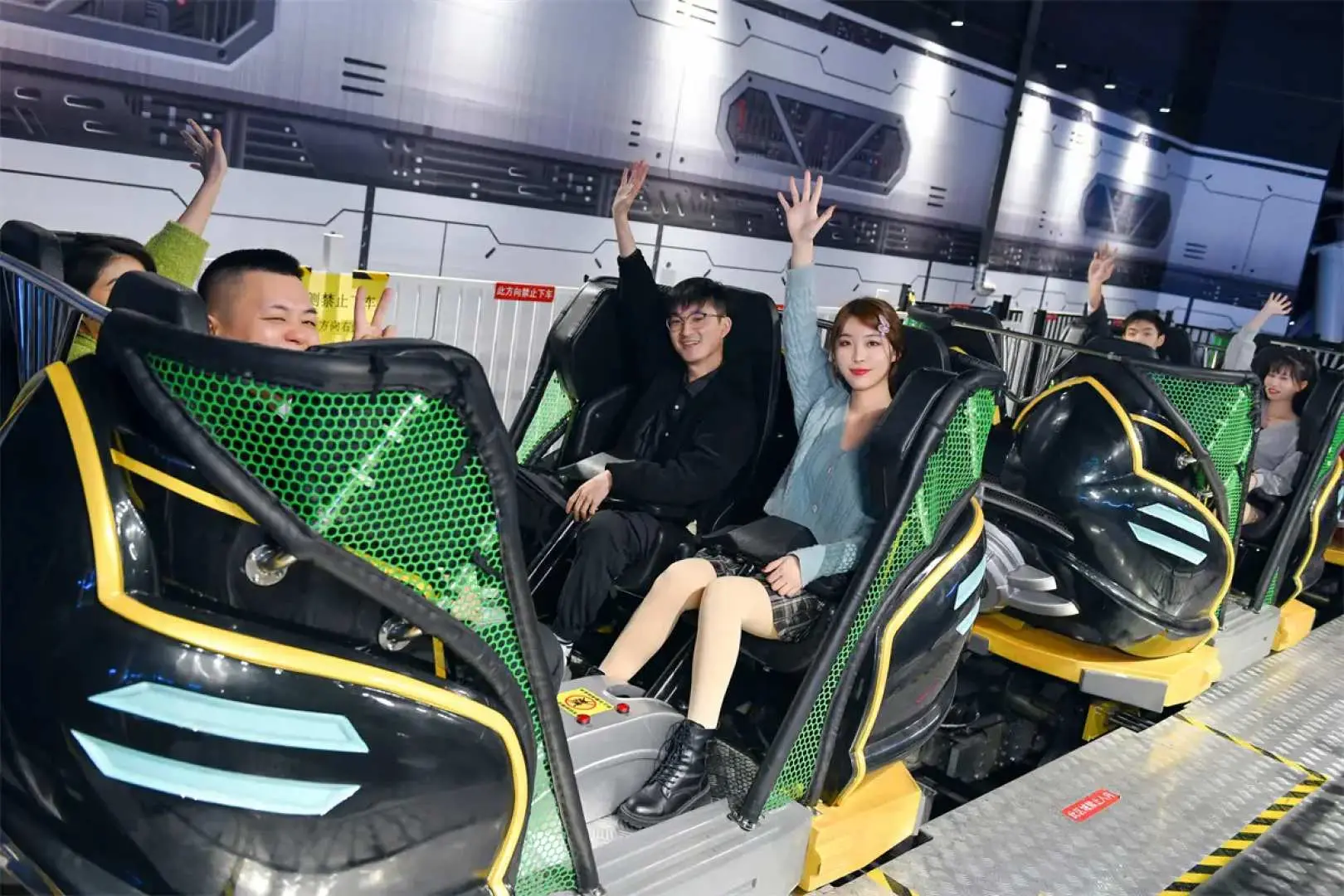 A group of excited visitors at Guangzhou Junming Happy World riding a thrilling indoor rollercoaster or simulation ride, raising their hands to enjoy the exhilarating speed.
