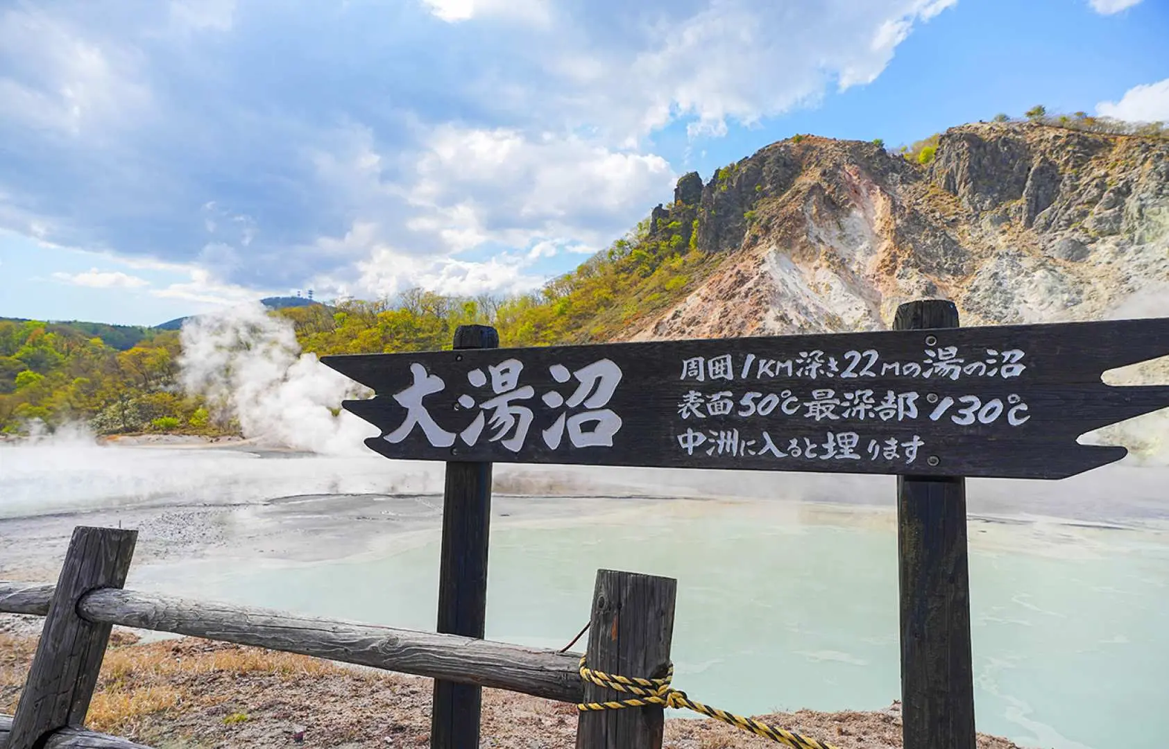 Noboribetsu Oyunuma sign, detailing the hot spring lake's size and temperature, with the steaming sulfur hot spring pond in the background, a famous Hell Valley attraction.