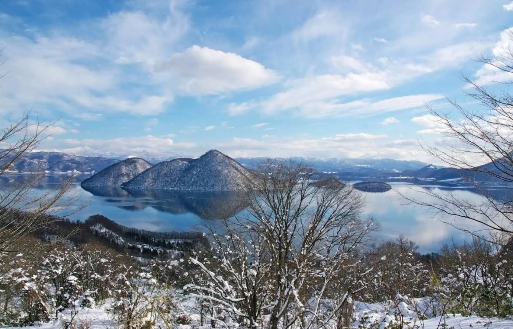 Expansive panoramic view of Lake Toya in winter, with its mirror-like surface reflecting snow-covered islands, adorned by blue sky and white clouds, showcasing Hokkaido's exquisite natural beauty.