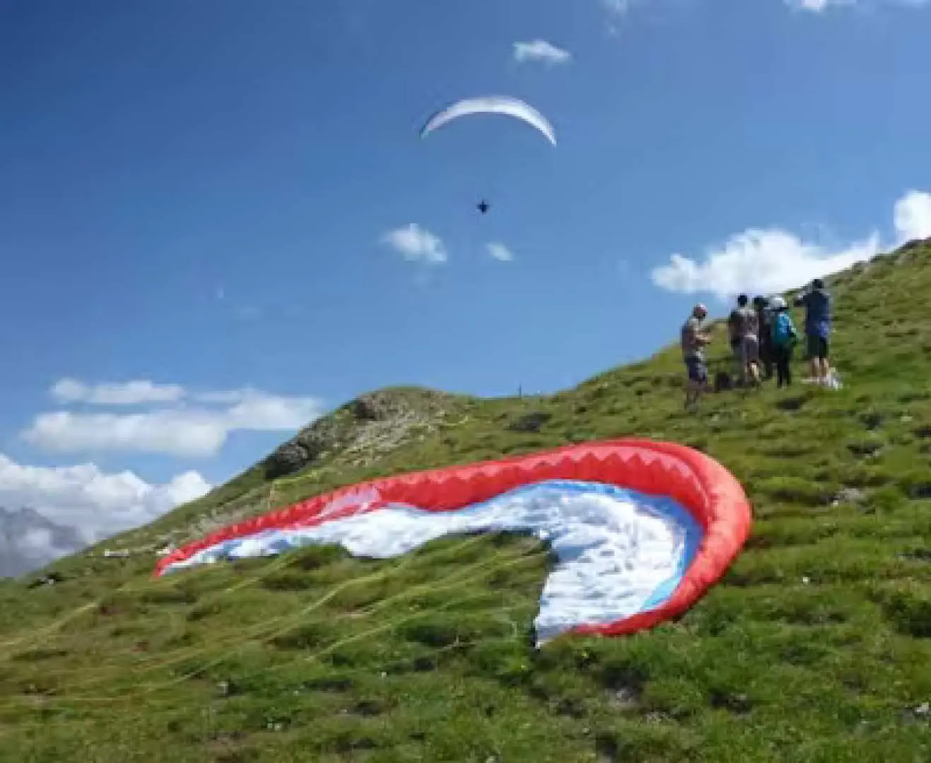スイスのクロスタースにある高山離陸地点で、赤、白、青のパラグライダーの翼が緑の斜面に広げられ、半日のスリリングな高空飛行の準備が整っています。遠くには別のパラグライダーが青空を飛んでいます。