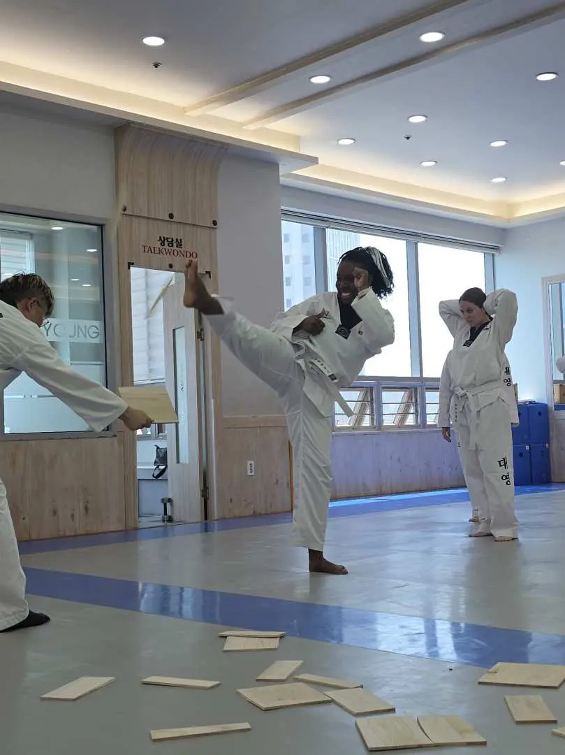 Một học viên tham gia trải nghiệm Taekwondo tại Busan thành công phá vỡ một tấm ván gỗ bằng cú đá cao mạnh mẽ, thể hiện kỹ năng và sự hướng dẫn an toàn.