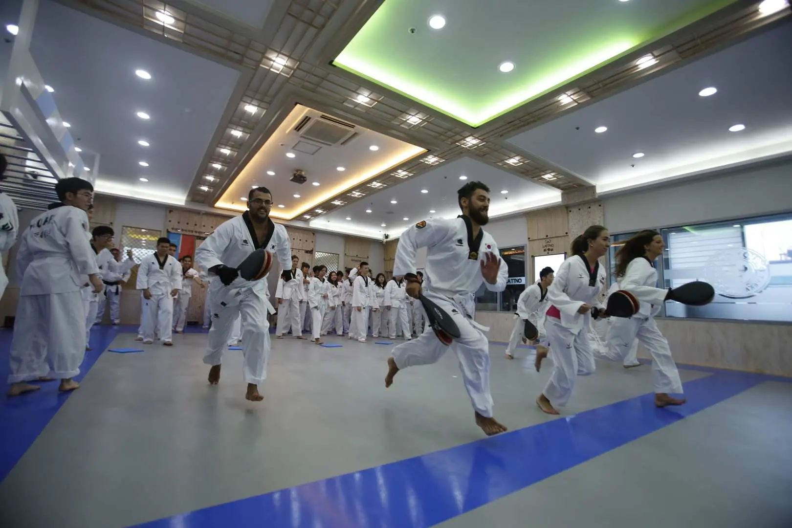 Các học viên nhiệt tình trong trang phục Taekwondo trắng chạy khắp sàn tập, cầm mục tiêu trong buổi khởi động năng động hoặc tập luyện cơ bản tại trải nghiệm võ thuật Busan.