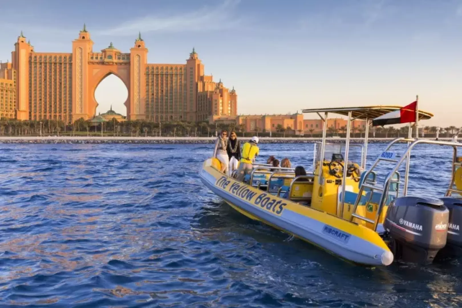 A yellow speedboat carrying passengers away from the camera, offering a clear view of the stunning Atlantis, The Palm resort from the open waters of Dubai's coastline.
