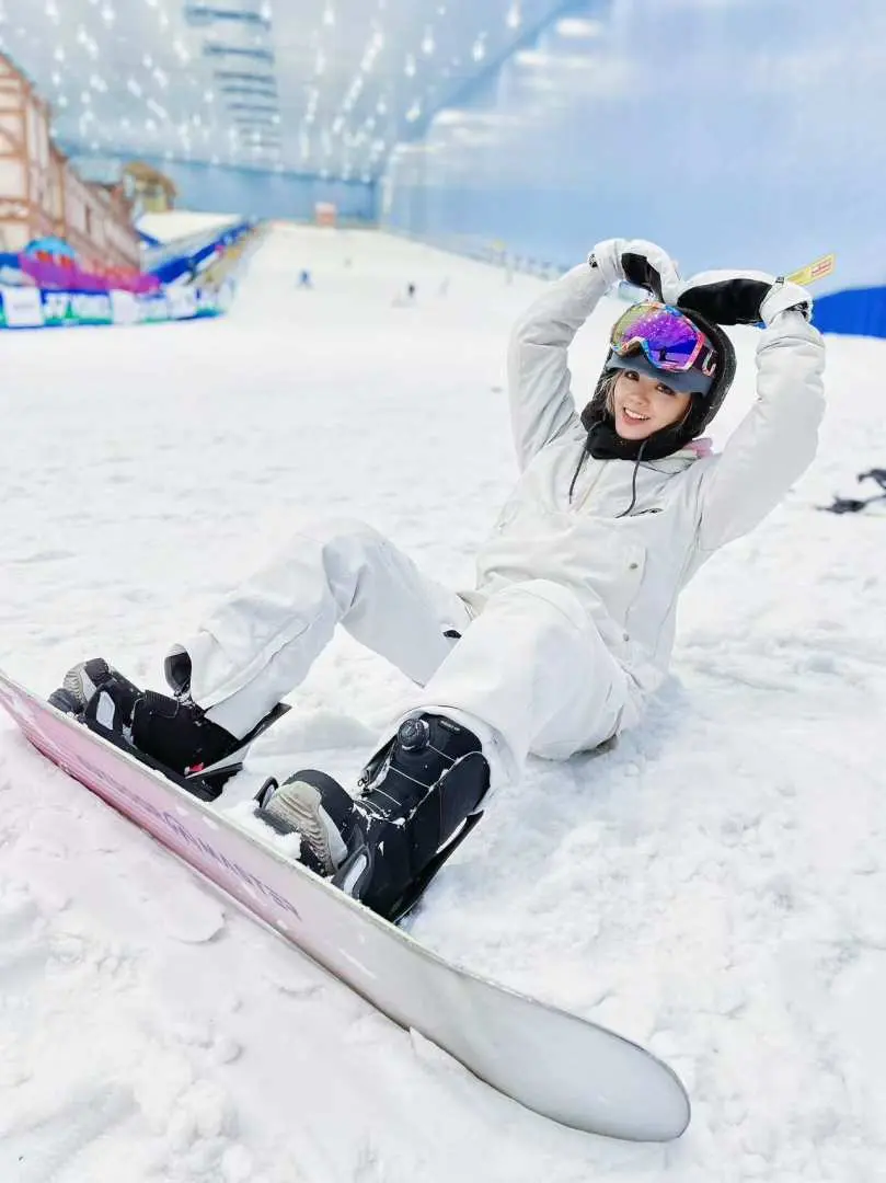 在深圳華發冰雪熱雪奇蹟滑雪場，穿白色滑雪服的女性單板滑雪者，面帶笑容坐在雪地上，享受滑雪後的輕鬆時刻。