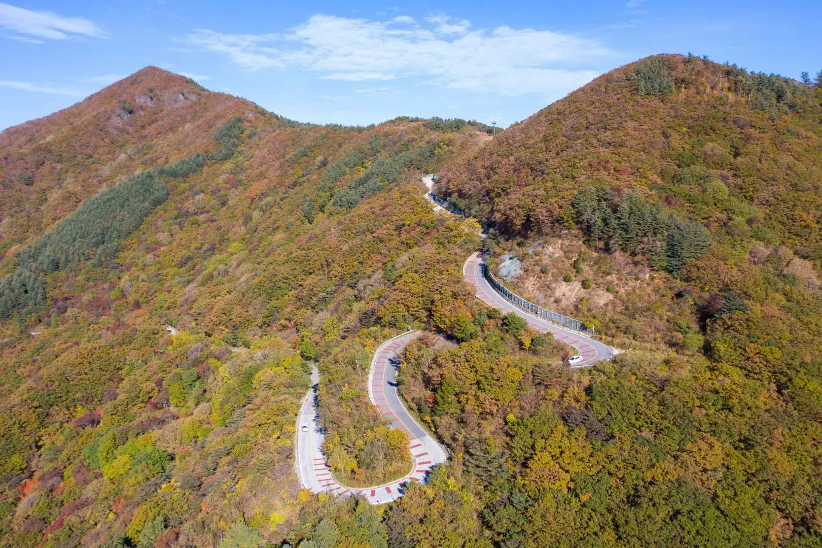 智异山秋季蜿蜒山路空拍，公路穿梭于层叠山峦间，两旁是染上秋色的树林，展现壮阔的山地美景。