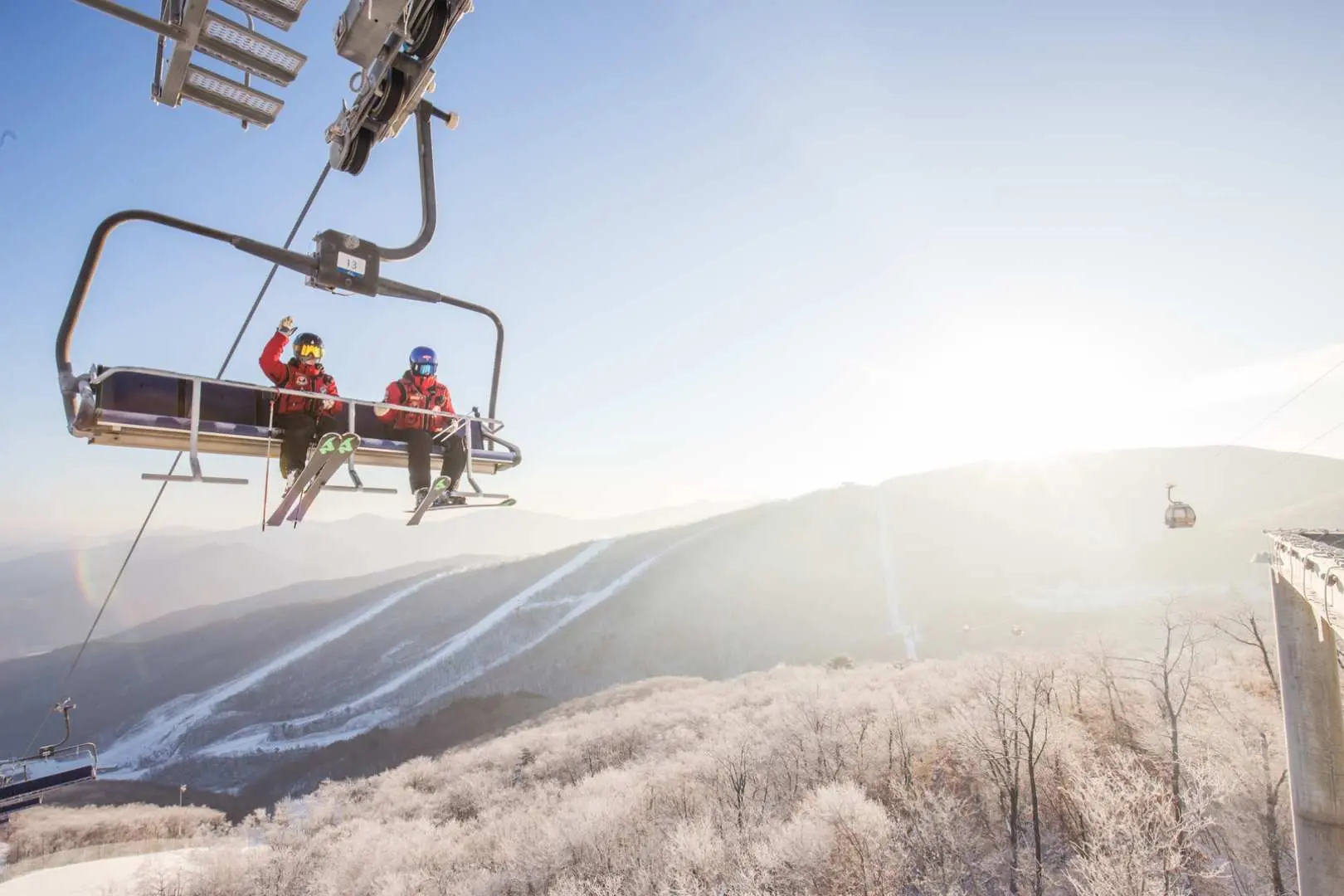 Two joyful skiers on a High1 Ski Resort chairlift give a thumbs-up, relishing the sunny, snow-covered mountain views and the exhilarating fun of skiing.