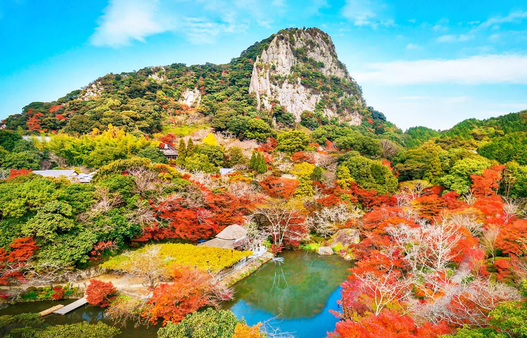 九州佐賀禦船山樂園的壯麗全景，禦船山岩壁聳立，湖水倒映著藍天，四周被火紅、金黃的秋楓環繞，呈現自然與人文之美。