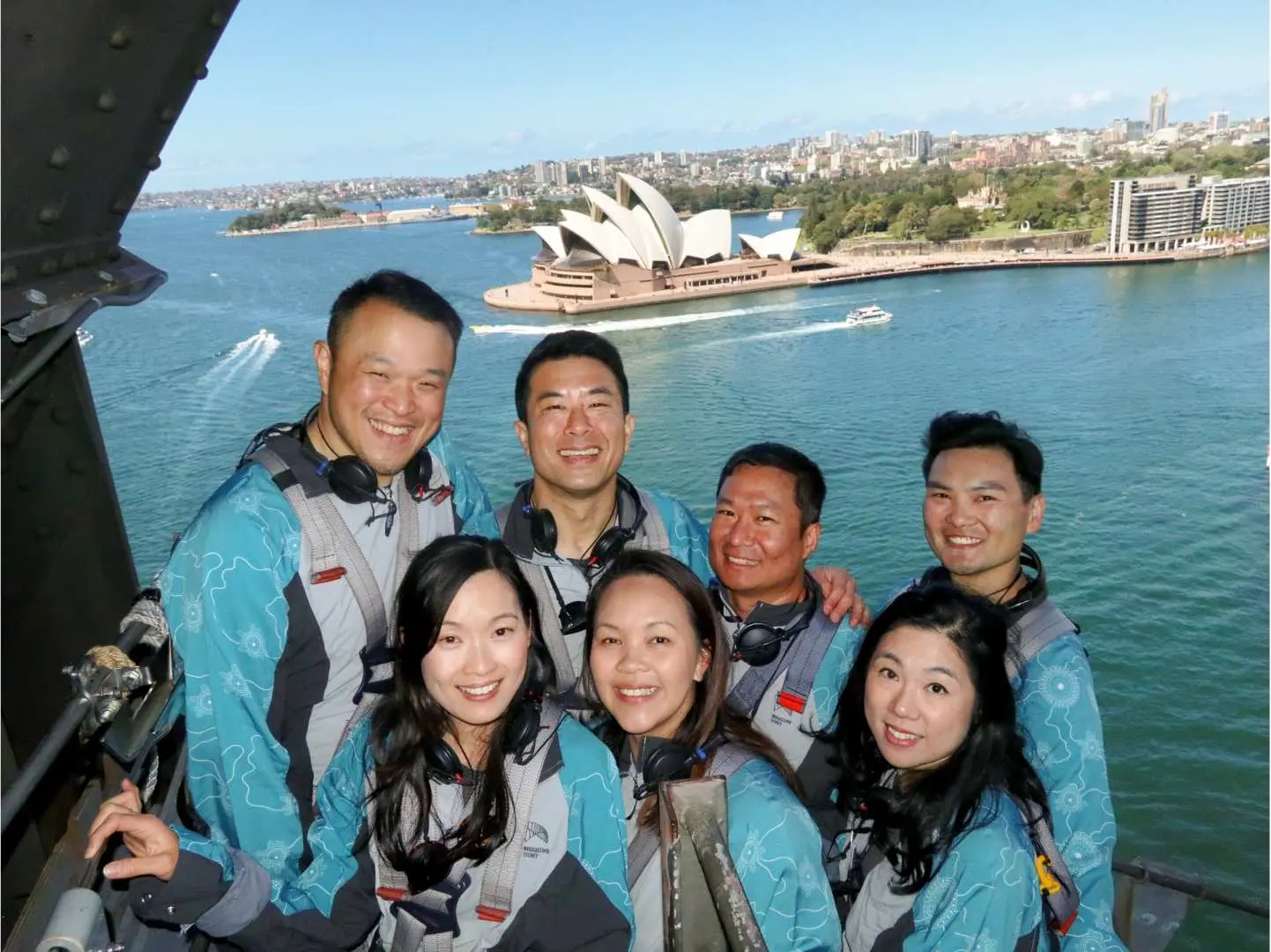 A group of smiling tourists in safety gear posing under the Sydney Harbour Bridge, with the iconic Sydney Opera House in the scenic background, showcasing a unique viewpoint.