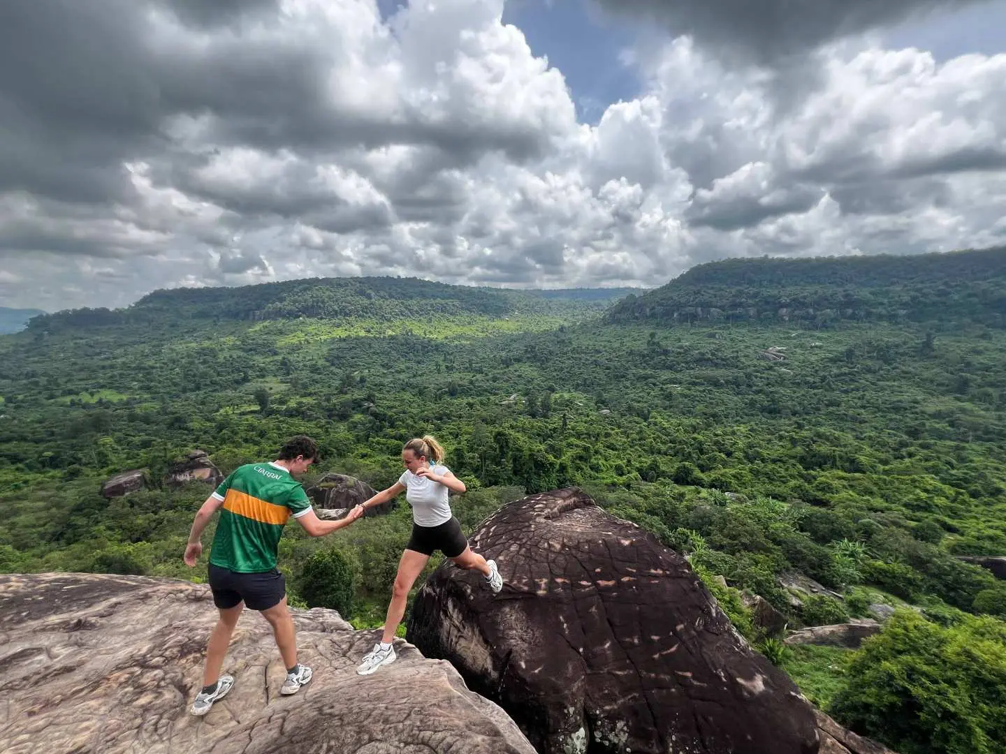 Couple enjoying the breathtaking panoramic views of the lush green landscapes and rolling hills from a rocky outcrop on Phnom Kulen, Cambodia.