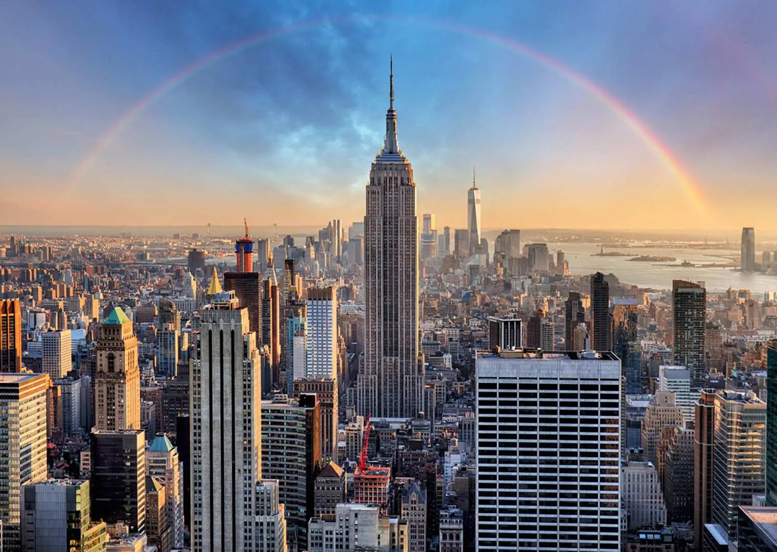 The iconic Empire State Building and New York City skyline bathed in golden sunset light, with a vibrant rainbow arching over the historic landmark.