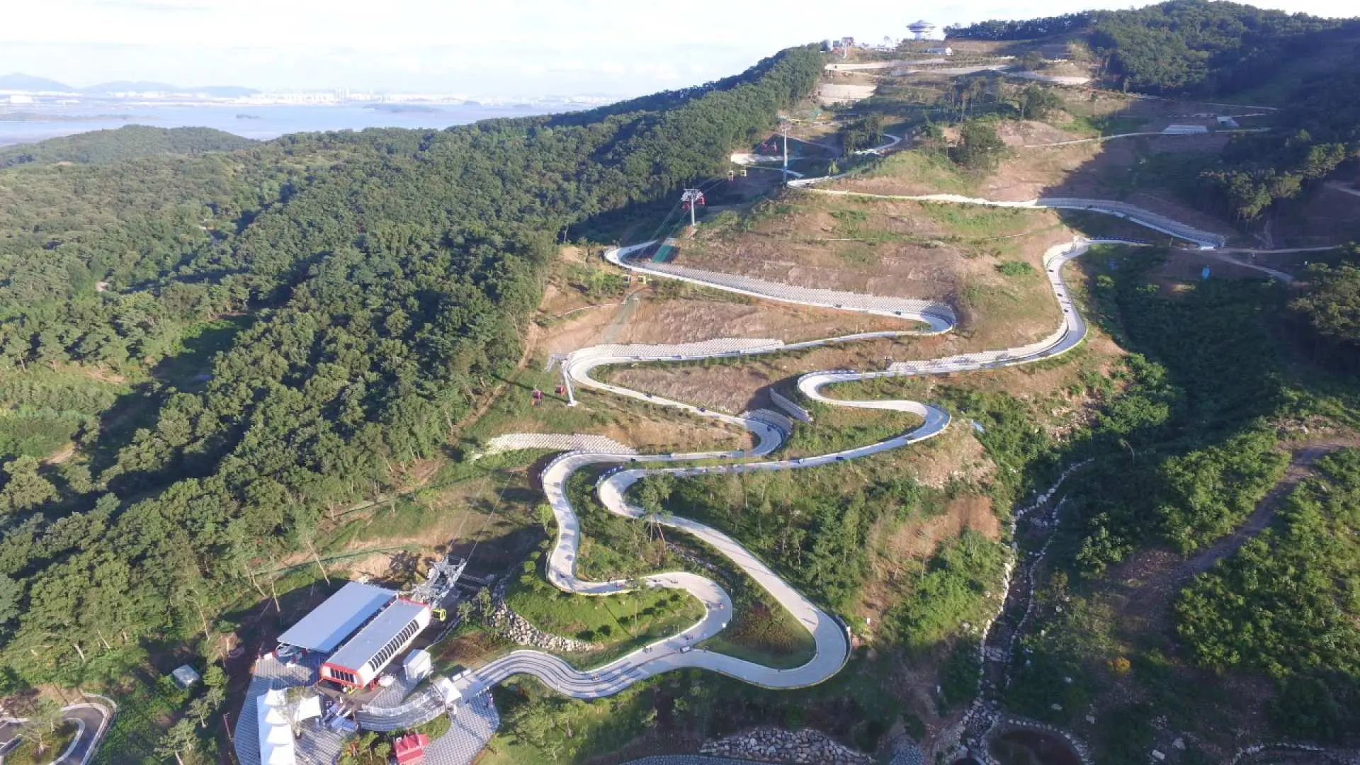 江华岛斜坡滑车蜿蜒赛道鸟瞰，从青翠山顶延伸至山脚，茂密树林环绕，远处海景尽收眼底，壮观。