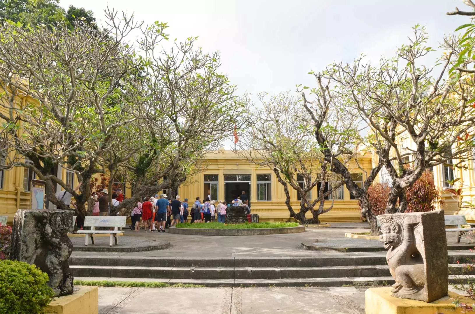 峴港佔婆雕刻博物館外觀，黃色法式殖民建築在綠樹環繞中顯得典雅，入口處有佔婆守護神獸石雕，遊客正步入其中。