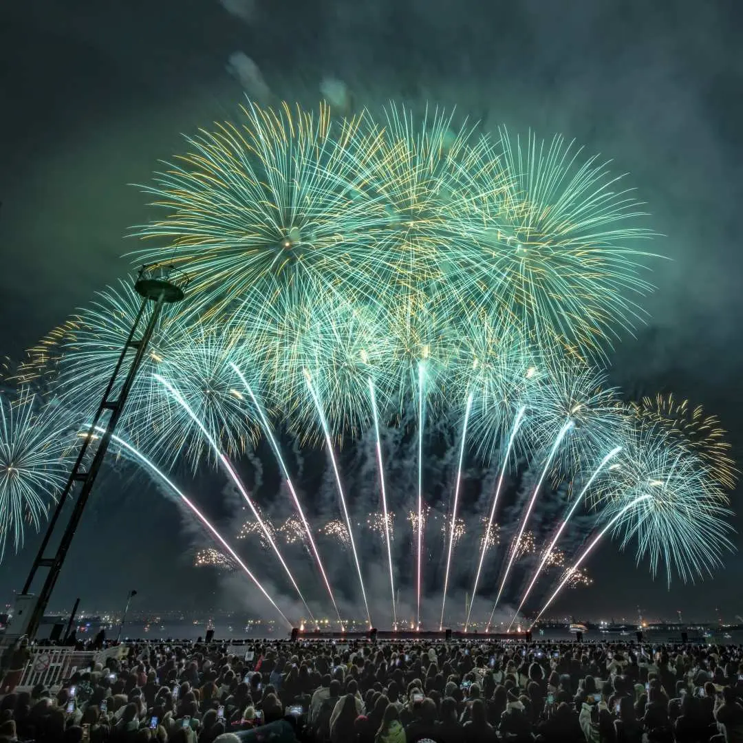 名古屋港いそがい花火劇場で、緑と青の花火が夜空を彩り、地上の光のショーと観客のシルエットが感動的な光景を作り出しています。