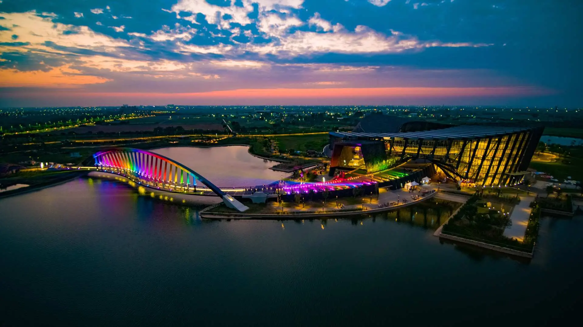 Aerial view of Chiayi Southern Branch of the National Palace Museum at dusk, with the rainbow bridge and museum building brilliantly illuminated, reflecting vibrant colors on the lake.
