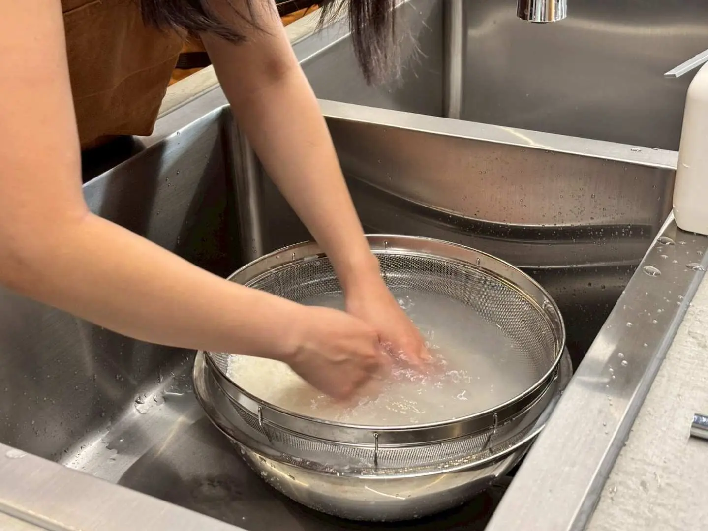 A participant in the Kagoshima traditional Japanese cooking class personally washes rice in a stainless steel sink, experiencing the foundational steps of preparing delicious firewood rice.