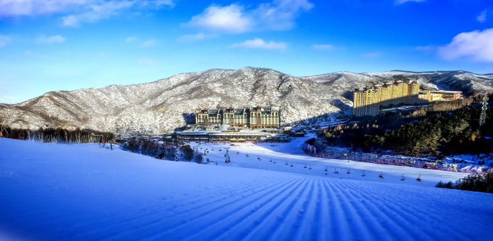 万龙滑雪场整齐压雪的雪道与宏伟的度假村建筑群，座落于崇礼山脉间，展现2022冬奥场地的世界级设施。