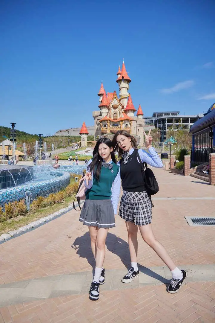 트렌디하고 다양한 K-POP 교복 스타일을 입은 두 친구가 'V' 사인을 하며 부산 롯데월드의 매혹적인 성 앞에서 즐겁게 포즈를 취하고 있습니다.