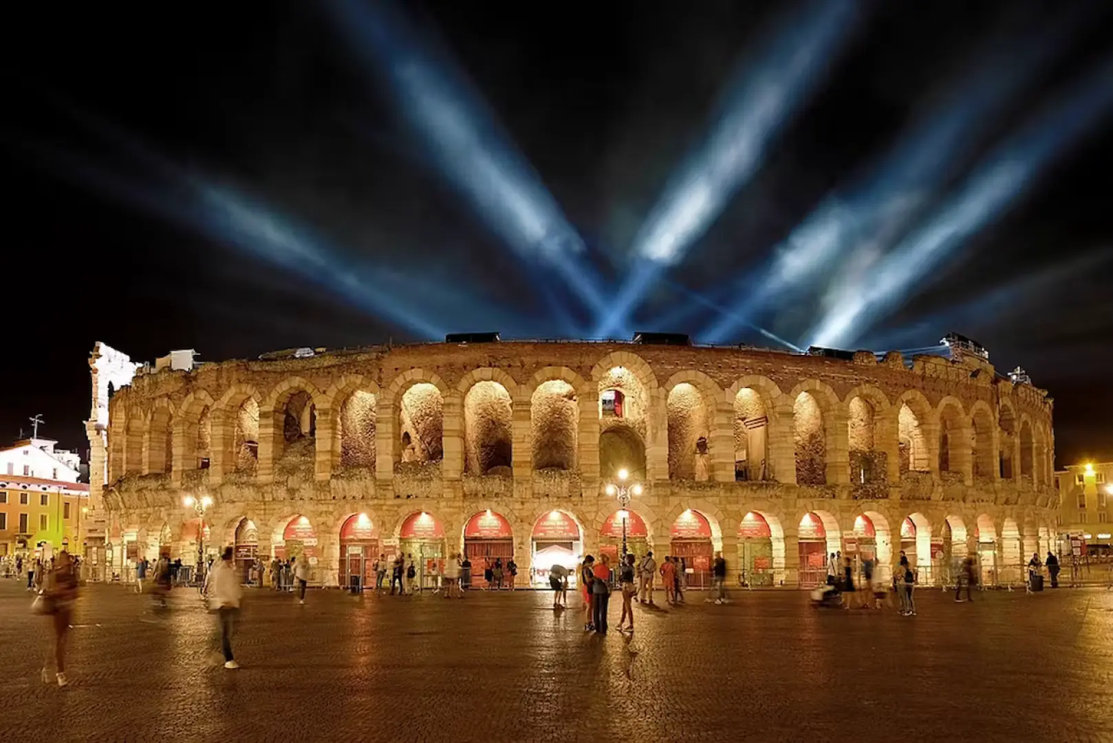 Captivating night view of the Verona Arena, illuminated by spotlights, showcasing ancient Roman ruins with visitors strolling in the piazza, offering free entry with the Verona Card.