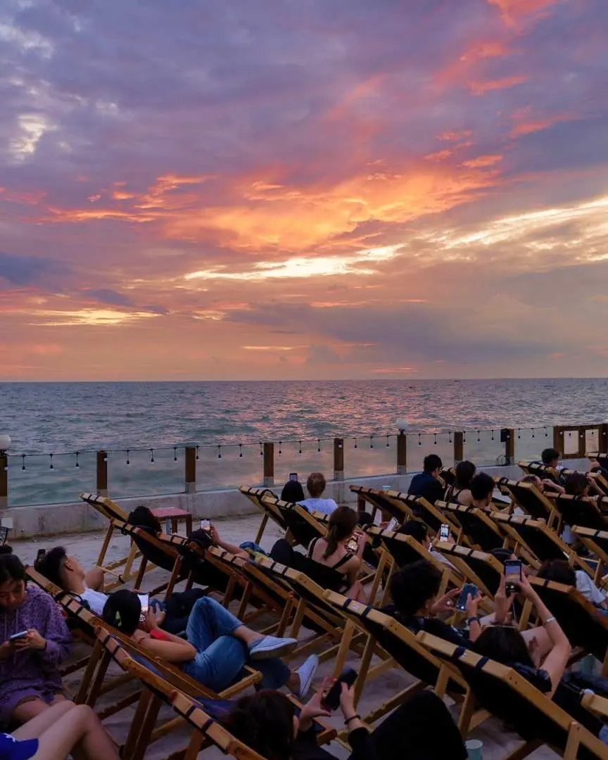 Visitors comfortably seated in beach chairs at Pattaya rooftop starlight cinema, enjoying the sparkling sea view at sunset, anticipating a unique movie experience after dark.