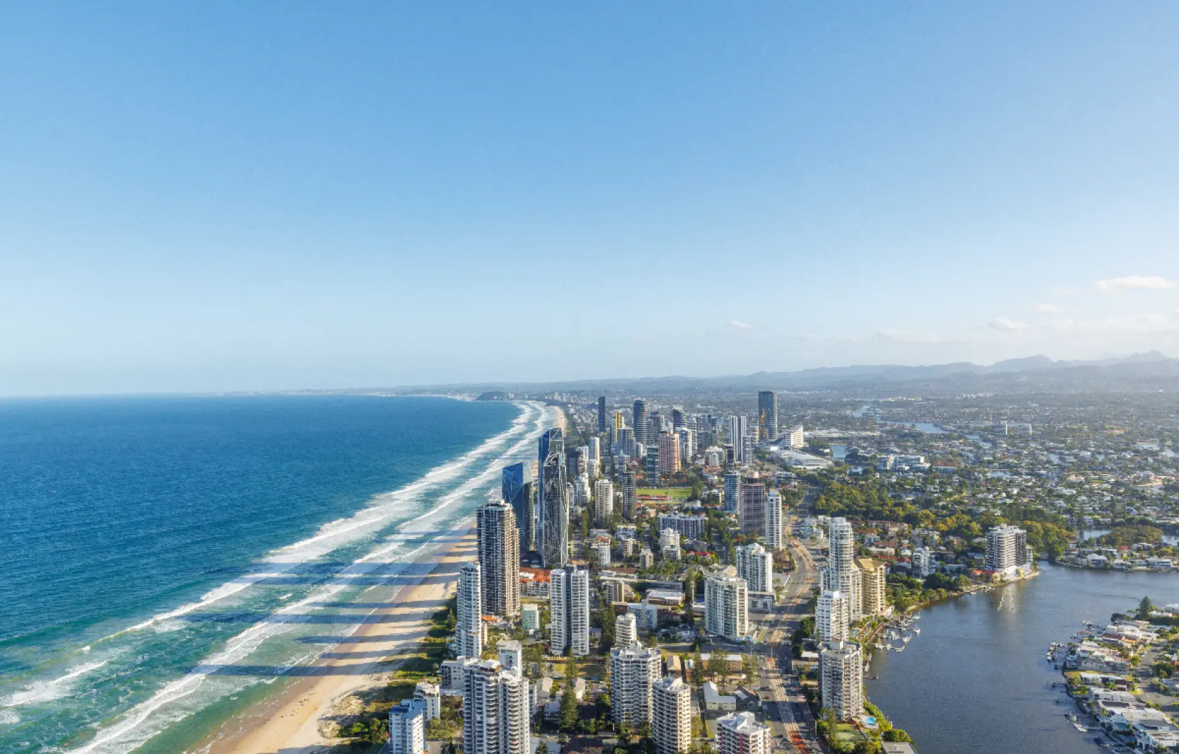 Stunning daytime aerial panorama of Gold Coast, Australia, featuring city skyscrapers stretching along golden beaches, with clear blue ocean waves under a bright, sunny sky.