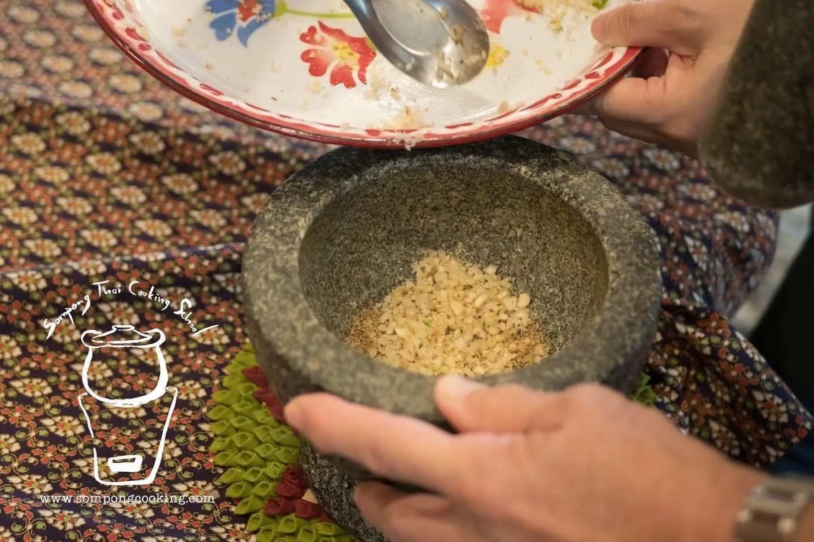 A close-up of hands grinding fresh spices with a traditional mortar and pestle at Sompong Thai Cooking Class in Bangkok, a key step in preparing authentic Thai cuisine.