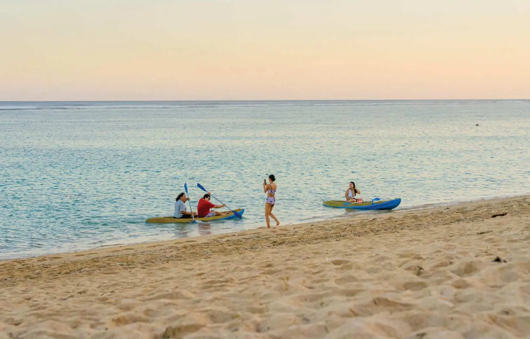 Three women joyfully kayaking in a yellow and blue tandem kayak in the clear blue ocean at Canna Nusa Dua, enjoying water sports and the tropical scenery.