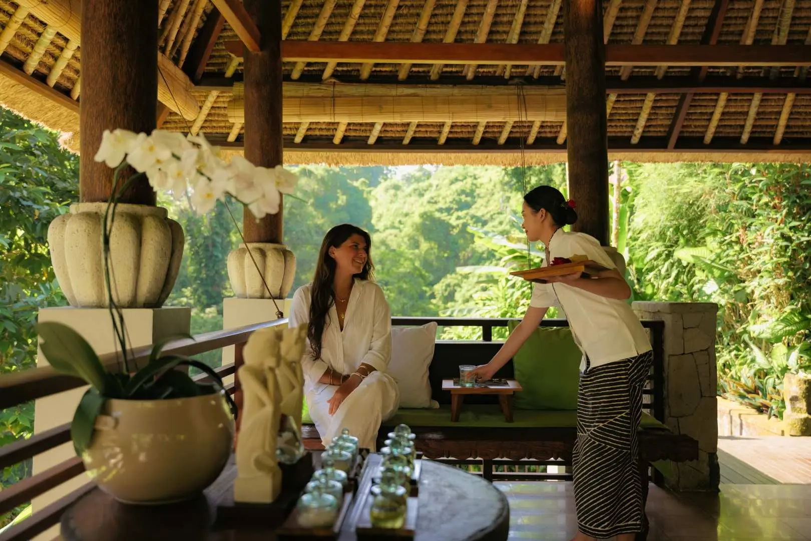 Spa staff at Maya Ubud Spa serving a guest in a robe with a welcome drink in an open-air lounge area, surrounded by the lush Bali jungle scenery.