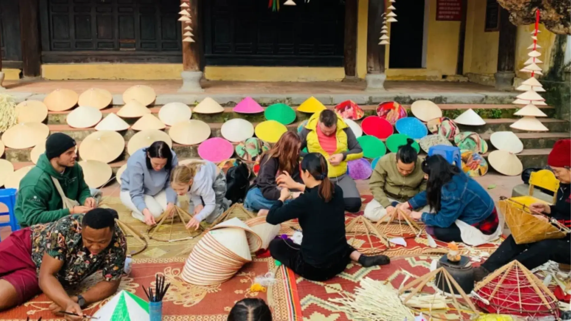 Du khách đang tích cực học cách đan nón lá truyền thống Việt Nam trong một xưởng thực hành tại làng Chương, xung quanh là nguyên liệu đầy màu sắc và sản phẩm hoàn chỉnh.