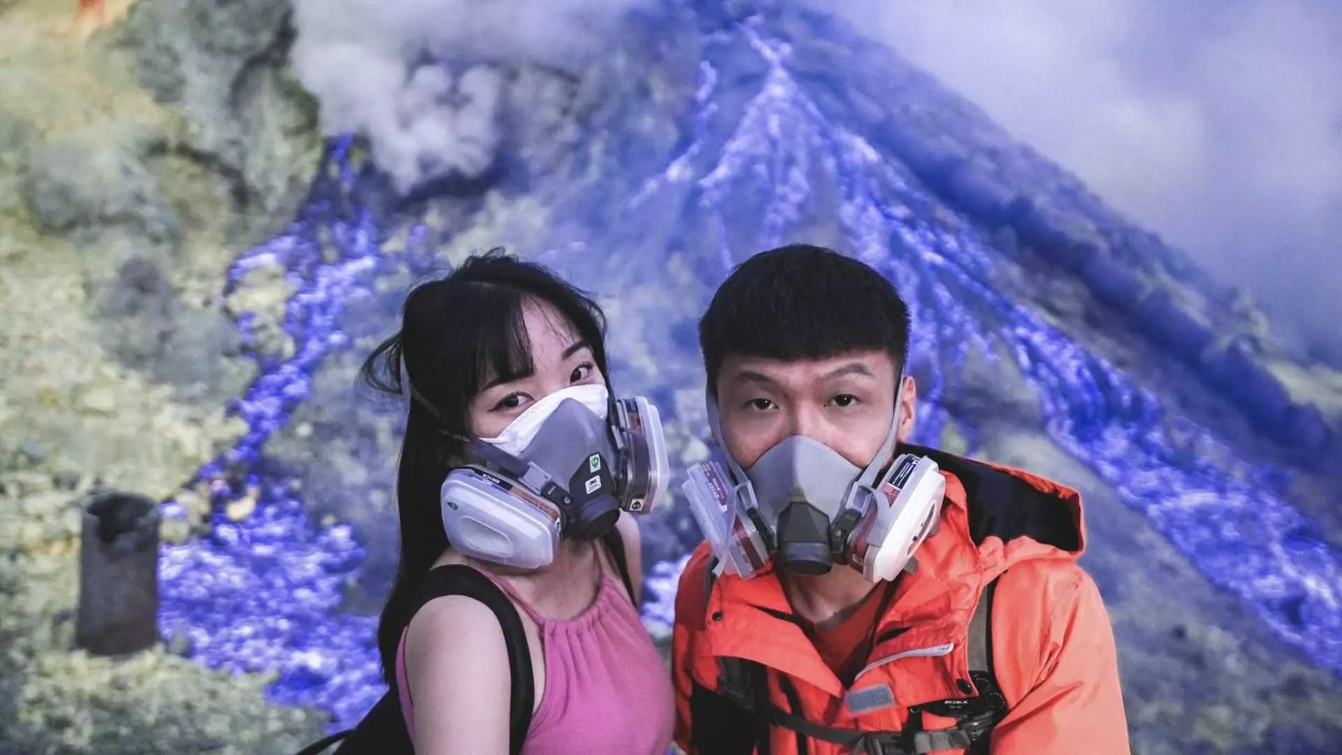 一對情侶戴著防毒面具，伊真火山口迷人的藍色火焰在身後若隱若現，準備體驗獨特的自然奇觀。