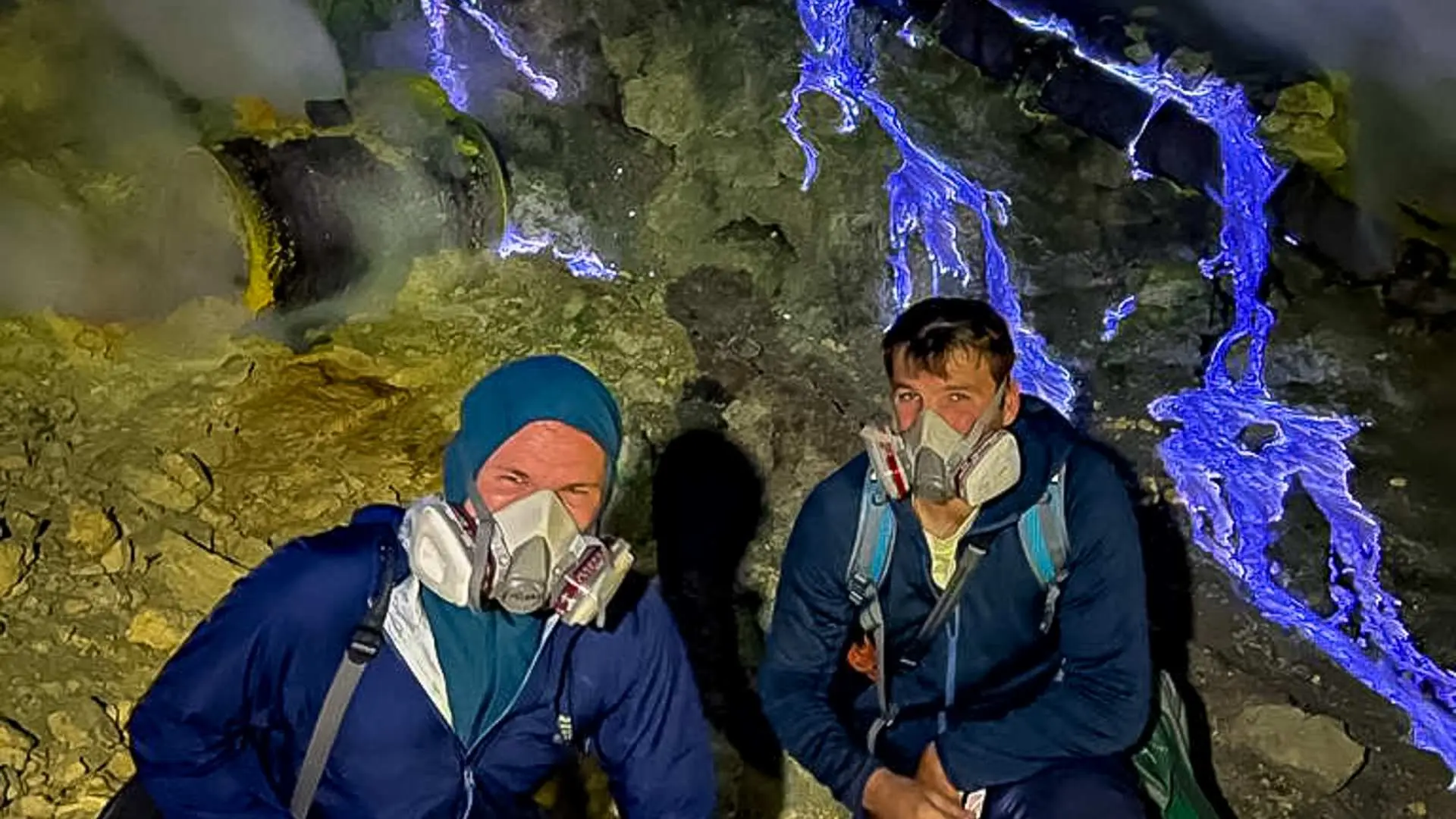 ガスマスクを着用した二人の旅行者が、夜間にイジェン火山の深部で電気のような青い炎を観察。神秘的で感動的な光景。