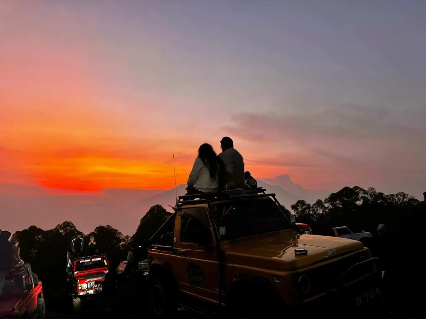 A couple sitting on a jeep roof, admiring a breathtaking sunset over Mount Batur in Bali, with the sky painted orange, creating a romantic atmosphere.