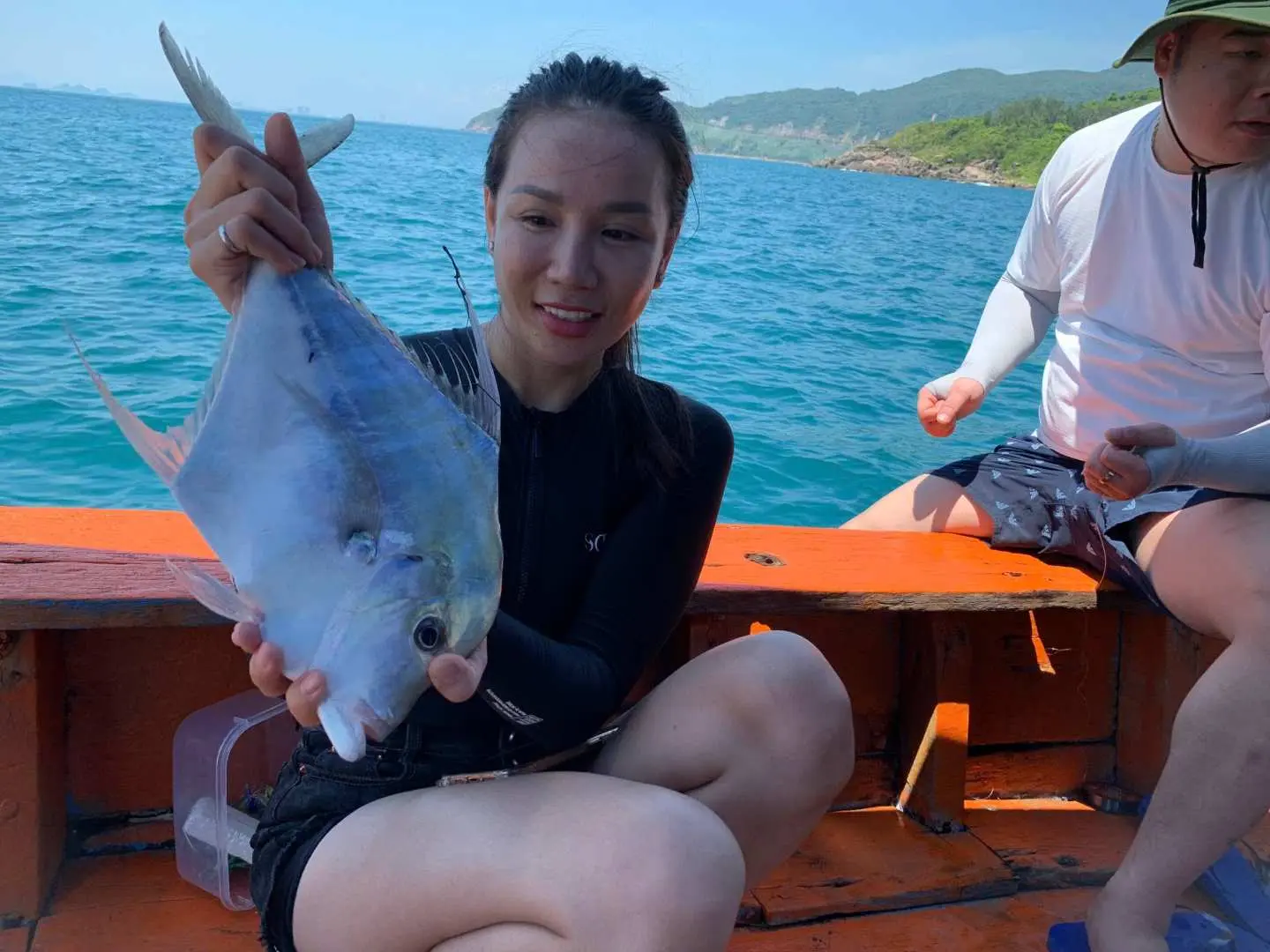 一位開朗女士在越南峴港山茶半島深海釣魚團中，自豪地展示她釣獲的銀色魚，藍色海域為背景。