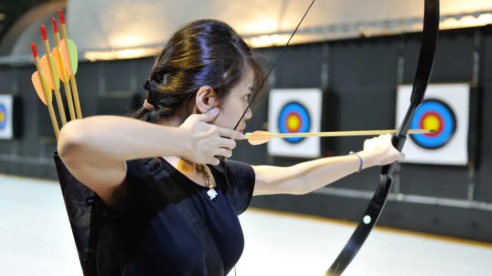 An archer at Taichung's Enlightenment Archery Range focusing on her stance indoors, equipped with a professional bow and arrows, practicing under expert guidance to master the art of archery.