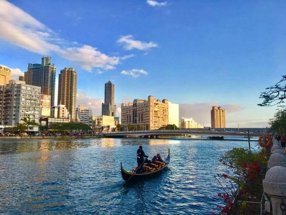 Tourists enjoying a gondola cruise on Kaohsiung's Love River under a clear blue sky, with towering buildings and bridges forming a magnificent city skyline along both banks.