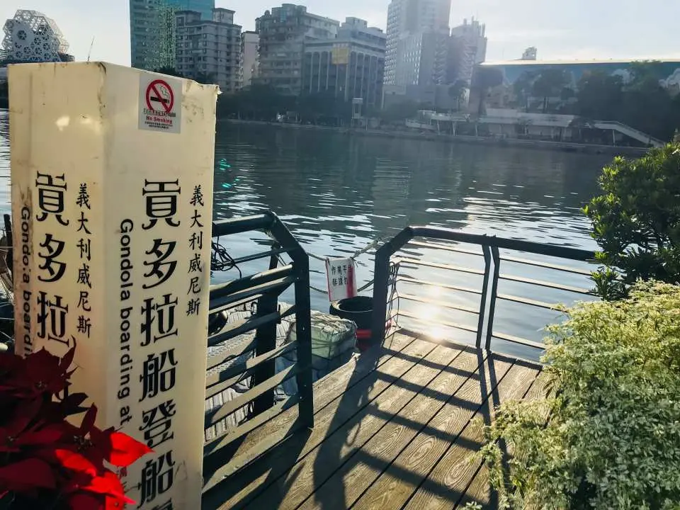 Close-up of the gondola boarding sign at Kaohsiung's Love River, with a sunny wooden pier and city buildings in the background, signaling the start of a romantic water journey.