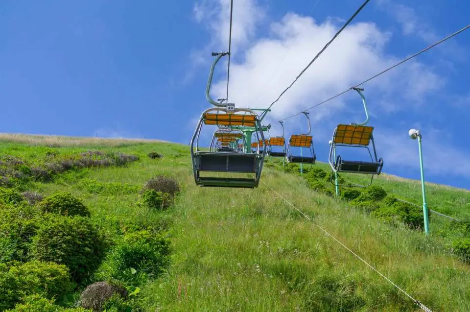 伊東の大室山または小室山の山頂へゆっくりと昇る観光客を乗せたリフト。青空と白い雲の下に広がる緑豊かな斜面、伊豆高原2日券で便利に観光。