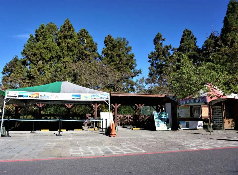 Outdoor transfer station or vendor area in Alishan National Forest Recreation Area, with shaded canopies under tall trees and a clear blue sky, for visitor convenience.