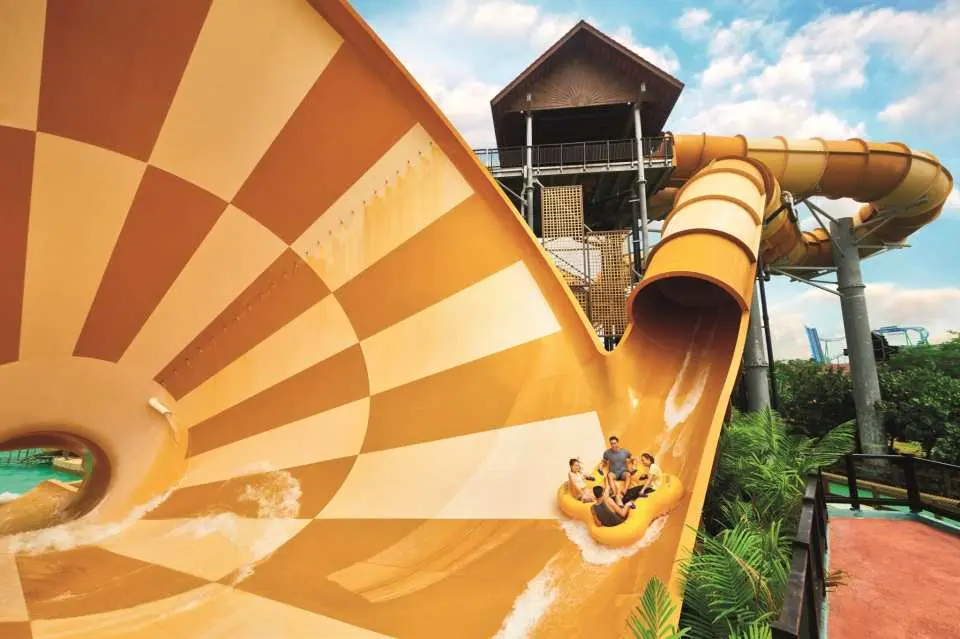 Thrill-seekers on an inflatable raft plunging down the large, checkered orange and white funnel slide at Desaru Coast Adventure Waterpark, captured from an overhead perspective.