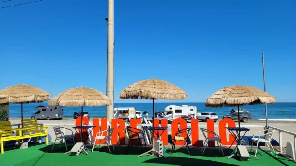 浦項新港のサーフィンリゾート、Surfholicの屋外休憩エリア。パラソルと大きなブランドロゴがあり、青い海とキャンピングカーを背景に、リラックスしたサーフィン体験を演出しています。
