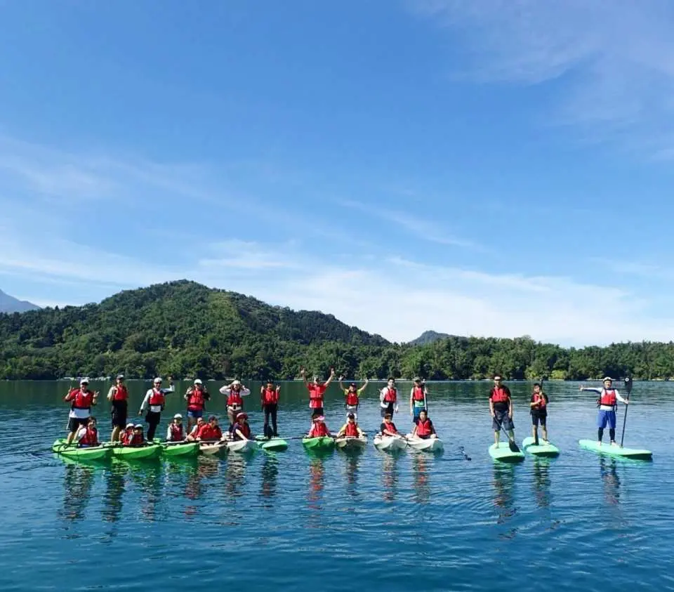 Large group photo of SUP paddleboarding and kayaking participants at Sun Moon Lake, happily posing amidst the blue sky and green waters, enjoying the fun of water activities.