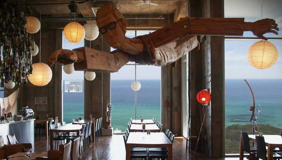 강릉 하슬라 아트월드 바다 카페. 커다란 목재 인체 조각상과 종이 등불이 매달려 있으며, 식사 공간에서는 손님들이 식사와 함께 탁 트인 바다 전망을 즐길 수 있습니다.